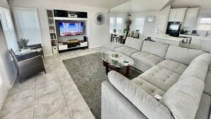A modern living room with a large flat-screen TV, grey sofas, a coffee table, and a dining area in the background. - Beachfront Paradise of Texas 4br 3 1/2 baths (Galveston)