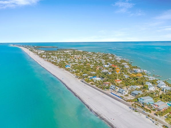 An aerial view of a coastal town with a long sandy beach, clear blue waters, and residential buildings.