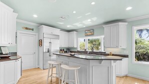 A kitchen with white cabinetry, a central island with stools, a refrigerator, and a window with a view of greenery. - Luxury 5BR Gulf-View Home with Private Pool, Spa & High-Tech Entertainment (Captiva)