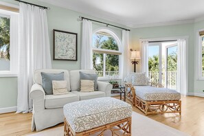 A room with a sofa, ottoman, wicker chair, and a table with a lamp.