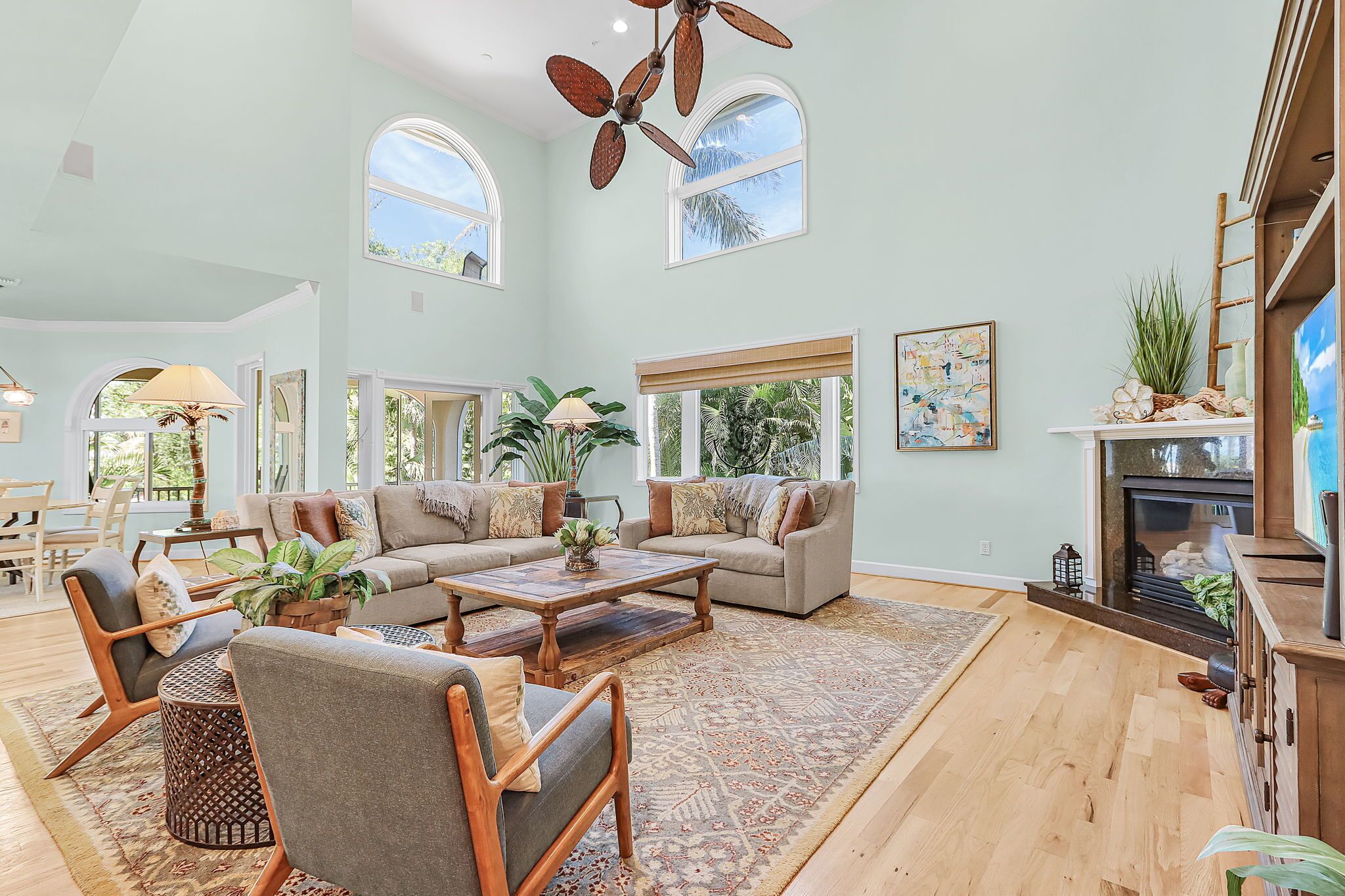 A spacious living room with a high ceiling, large windows, a fireplace, and a ceiling fan.