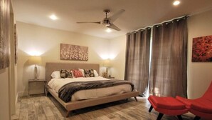 A bedroom with a bed, a ceiling fan, two bedside lamps, a red chair, and a window with curtains. - Modern Waterfront Home with Hot Tub, Dock, Kayaks (New Port Richey)