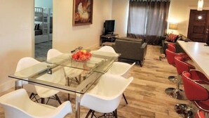 A modern dining area with a glass table and white chairs, a red bar area with stools, and a living room in the background. - Modern Waterfront Home with Hot Tub, Dock, Kayaks (New Port Richey)