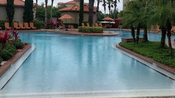 A swimming pool with palm trees and lounge chairs, surrounded by buildings with red-tiled roofs.