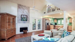 A spacious living room with a fireplace, a dining area with a white table, and a kitchen in the background.