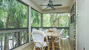 A covered outdoor dining area with a wooden table and wicker chairs, surrounded by large windows offering a view of tropical foliage.