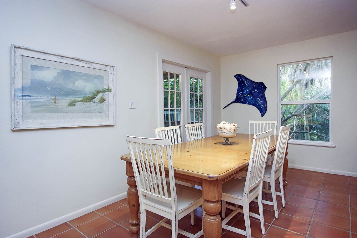 A dining room with a wooden table and white chairs, a painting on the wall, and a window with a view of trees.