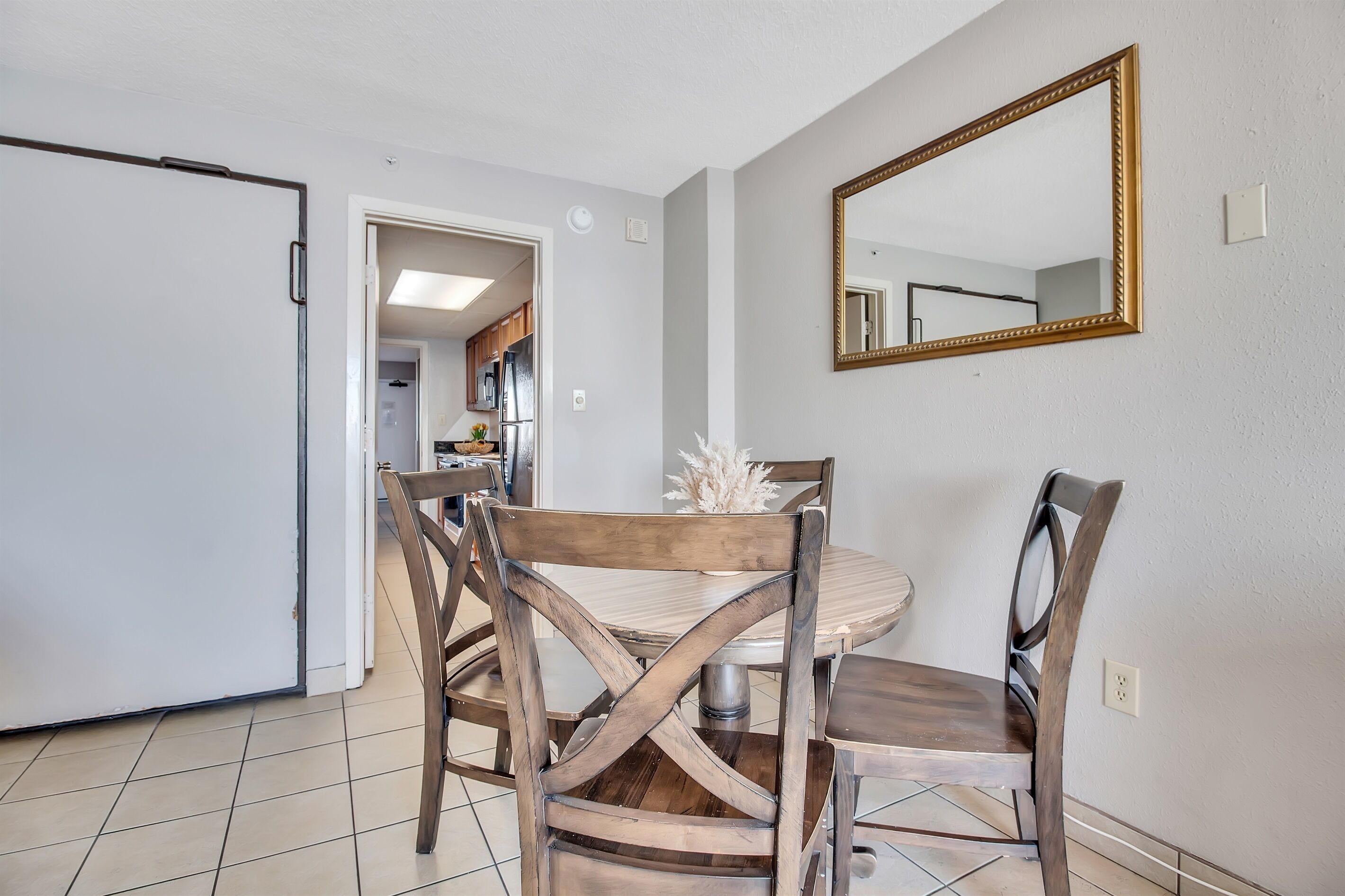 Un comedor con una mesa redonda y sillas de madera, un espejo en la pared y una cocina visible a través de una puerta abierta.