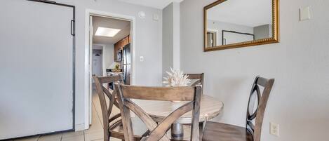 Un comedor con una mesa redonda y sillas de madera, un espejo en la pared y una cocina visible a través de una puerta abierta.