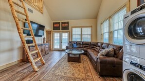 A spacious living room with a large brown leather sofa, a glass coffee table, a wooden ladder, a washing machine, and a TV mounted on the wall. - Lake Travis Waterfront with Resort Style Pool and Million Dollar Views. (Marble Falls)