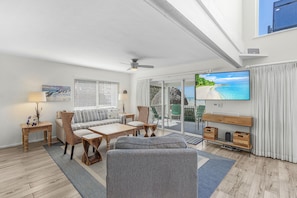 A spacious living room with a large TV, comfortable seating, and a balcony with a view of the ocean.