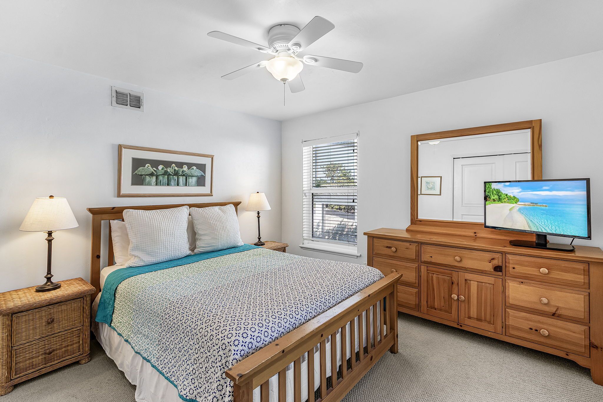 A bedroom with a bed, bedside lamps, a ceiling fan, a dresser, a TV, and a window with blinds.