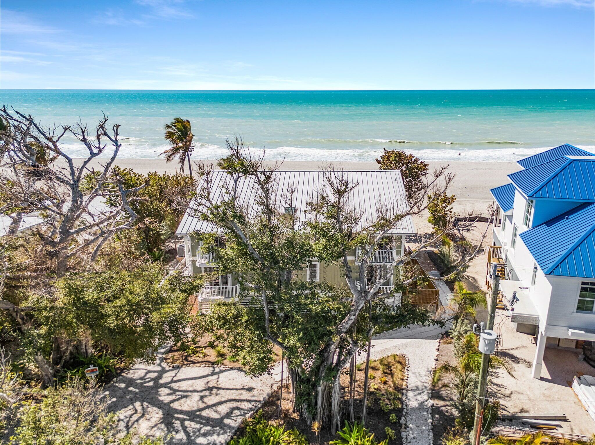Une propriété située en bord de mer, avec un bâtiment blanc et une structure surmontée d’un toit bleu, entourée d’arbres et bordée d’une plage de sable.