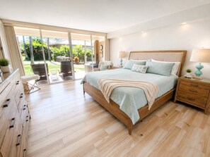 A bedroom with a large bed, wooden nightstands, a dresser, and a sliding glass door leading to a patio with outdoor furniture.