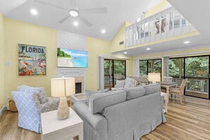 Amplia sala de estar con ventilador de techo, televisor de pantalla plana, chimenea y ventana grande con vistas a la vegetación.
