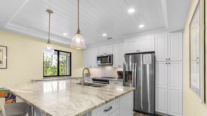 Una cocina con armarios blancos, encimera de mármol, electrodomésticos de acero inoxidable e iluminación colgante.