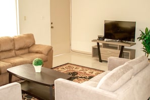 A living room with a beige sofa, a coffee table with a plant, a TV on a wooden stand, and a patterned rug.