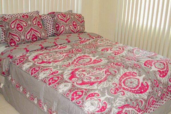A bed with a patterned bedspread and pillows, a window with blinds, and a plain wall.