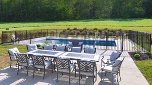 An outdoor patio with a pool, surrounded by a fence, and a grassy area in the background.