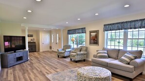 A spacious living room with hardwood flooring, a grey entertainment unit, a beige sofa with striped cushions, two patterned armchairs, and a round ottoman.