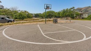 An outdoor basketball court with a hoop, surrounded by a landscaped area with trees and shrubs. - Oceanfront at Ships Watch! (Duck)