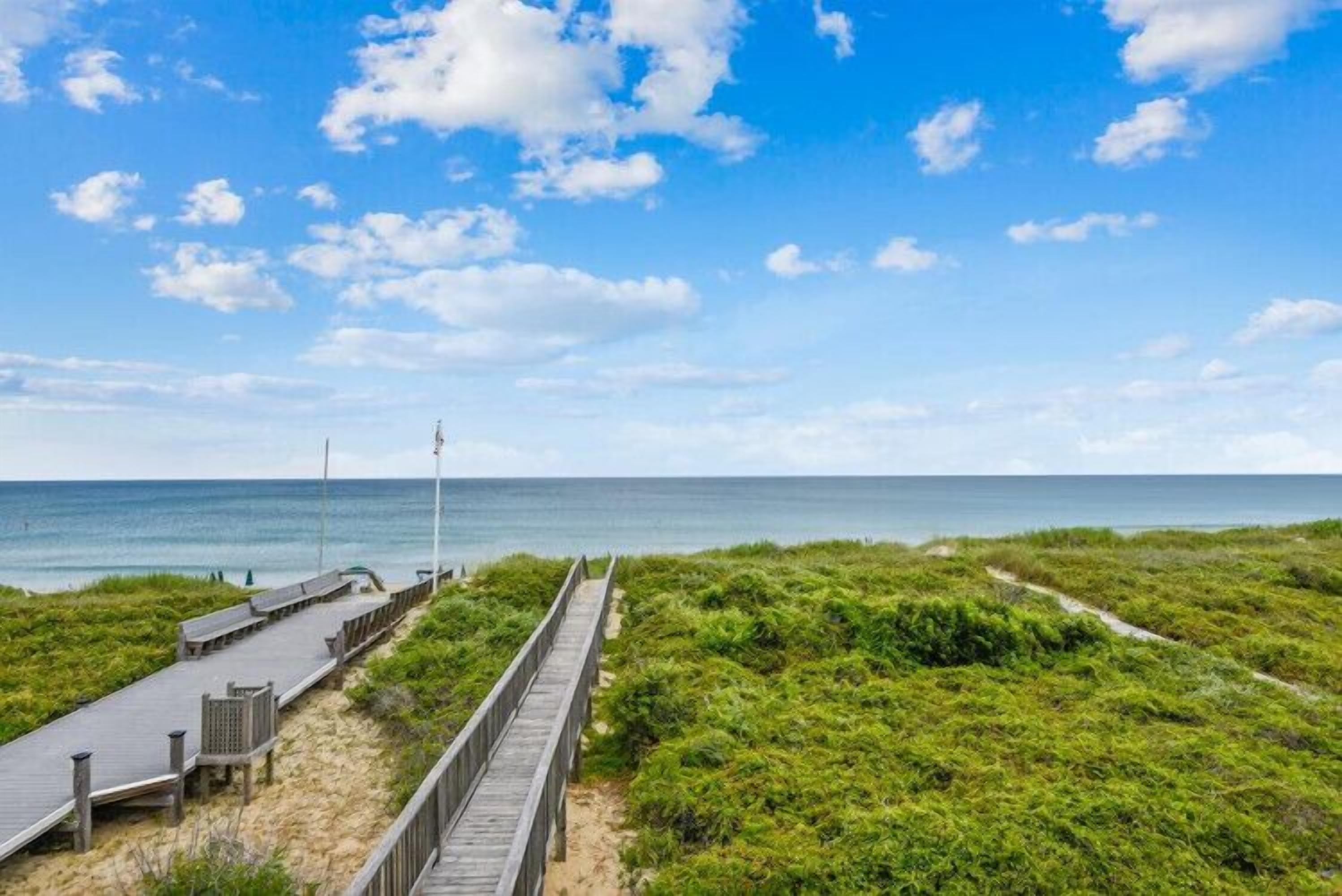 A coastal boardwalk with a wooden walkway, a viewing platform, and a grassy area leading to the sea.
