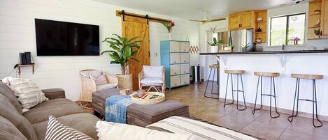 A modern living room with a large sofa, a coffee table, and a TV. There is a kitchen area with a bar and stools.