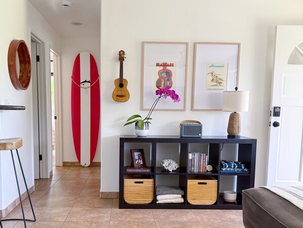 A hallway with a surfboard, a guitar, framed pictures, a plant, and a console table with books and decorative items.