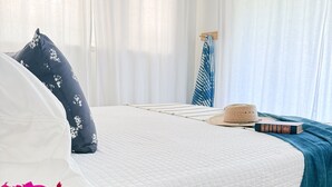 Ein Schlafzimmer mit Bett, weißen Vorhängen, einem blau-weiß gestreiften Handtuch, einem Hut und einem Buch auf dem Bett.