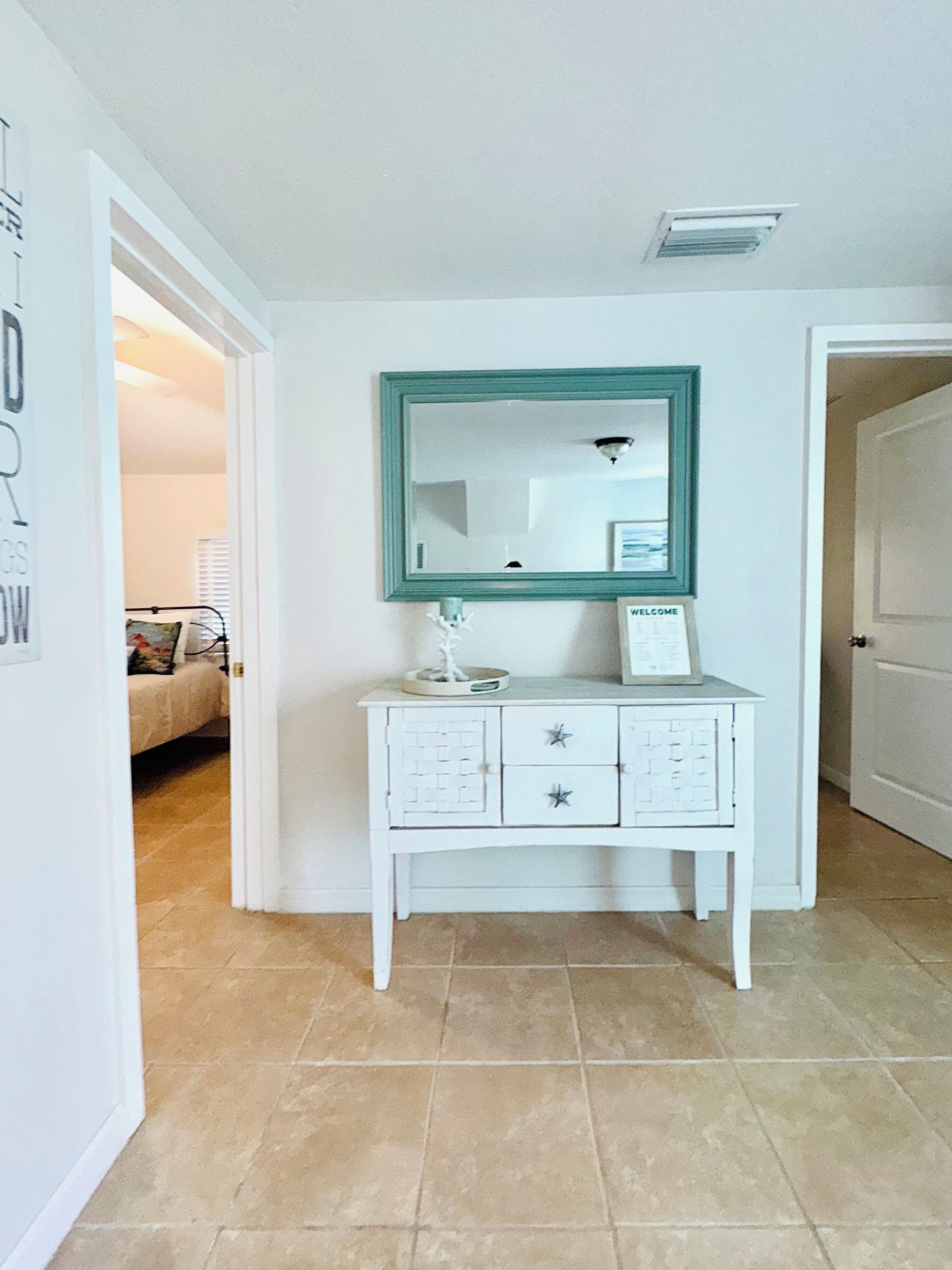 A white vanity with a teal-framed mirror, a small white cabinet, and a welcome sign.