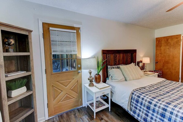 Un dormitorio con puerta de madera, una mesita de noche con lámpara, una cama con manta escocesa y una estantería con objetos decorativos.