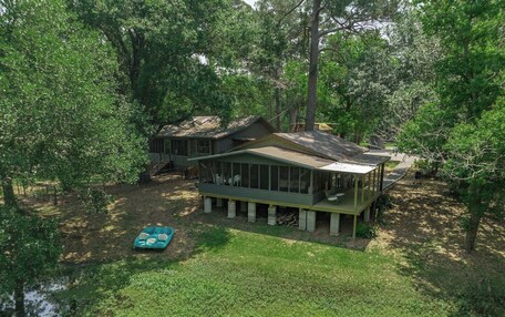 A two-story house with a large deck and a covered porch, surrounded by dense trees and a small pond.