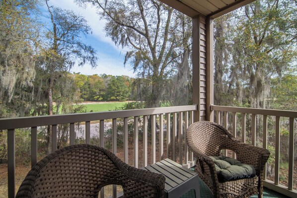 A porch with wicker chairs and a table, overlooking a wooded area with a view of a body of water.