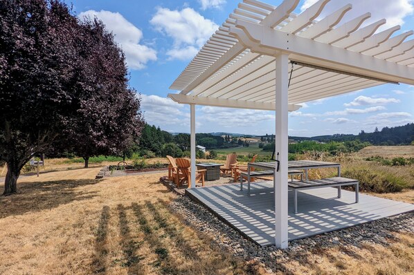 A covered outdoor seating area with a pergola, wooden chairs, and a table, overlooking a scenic landscape.