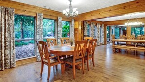 A spacious dining area with wooden tables and chairs, large windows with curtains, and a chandelier. - 8 acres of fun & relaxation in a beautiful Rock Cottage with a pool and hot tub (Tomball)