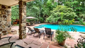 A stone patio with a pool, surrounded by lush greenery and a wooden deck. - 8 acres of fun & relaxation in a beautiful Rock Cottage with a pool and hot tub (Tomball)