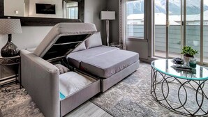 Ein modernes Wohnzimmer mit Schlafsofa, Glastisch, Lampe und Bergblick durch große Fenster.