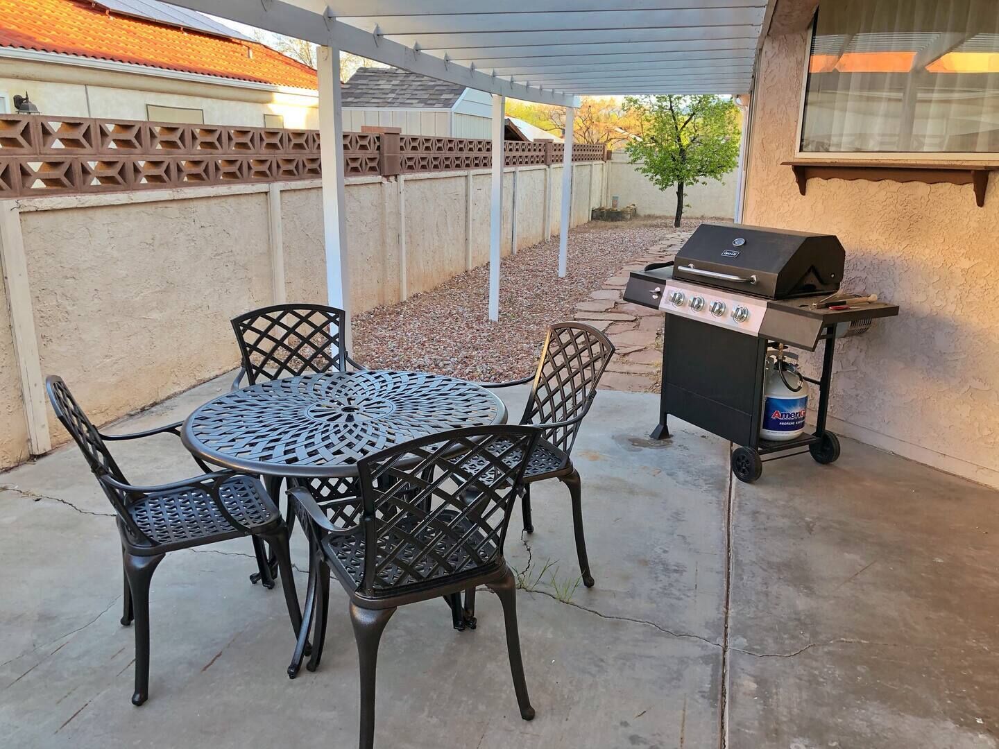 Une terrasse extérieure avec une table ronde en métal et quatre chaises, un barbecue et une bouteille de propane.