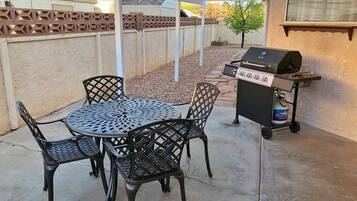Une terrasse extérieure avec une table ronde en métal et quatre chaises, un barbecue et une bouteille de propane.