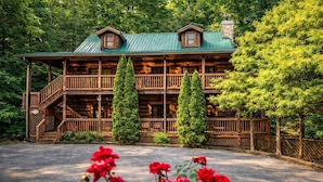A two-story log cabin with a green metal roof, wooden balconies, and a stone chimney.