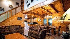 A cozy cabin interior with wooden walls, a staircase, a living room with a sofa and coffee table, a kitchen area, and a dining table.