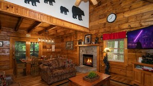 A log cabin interior with a fireplace, wooden furniture, a clock, and a TV displaying a cityscape.
