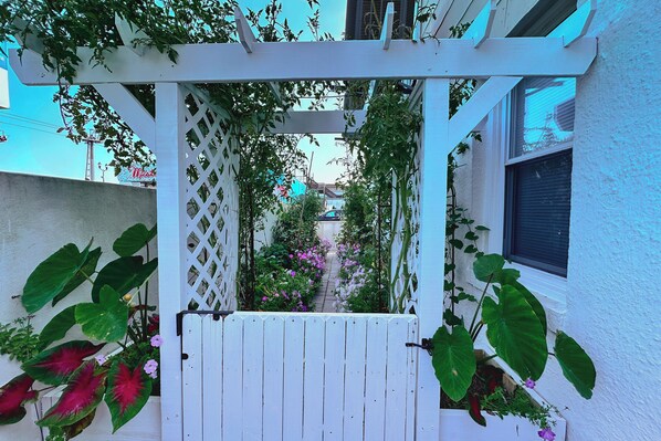 Um portão de madeira branca com design treliçado, rodeado por plantas verdes exuberantes e flores vibrantes.