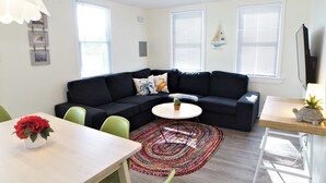 Una sala de estar moderna con un sofá seccional negro, una mesa redonda de centro y una mesa de comedor con sillas. Hay una alfombra colorida en el suelo y una decoración con forma de velero en la pared.
