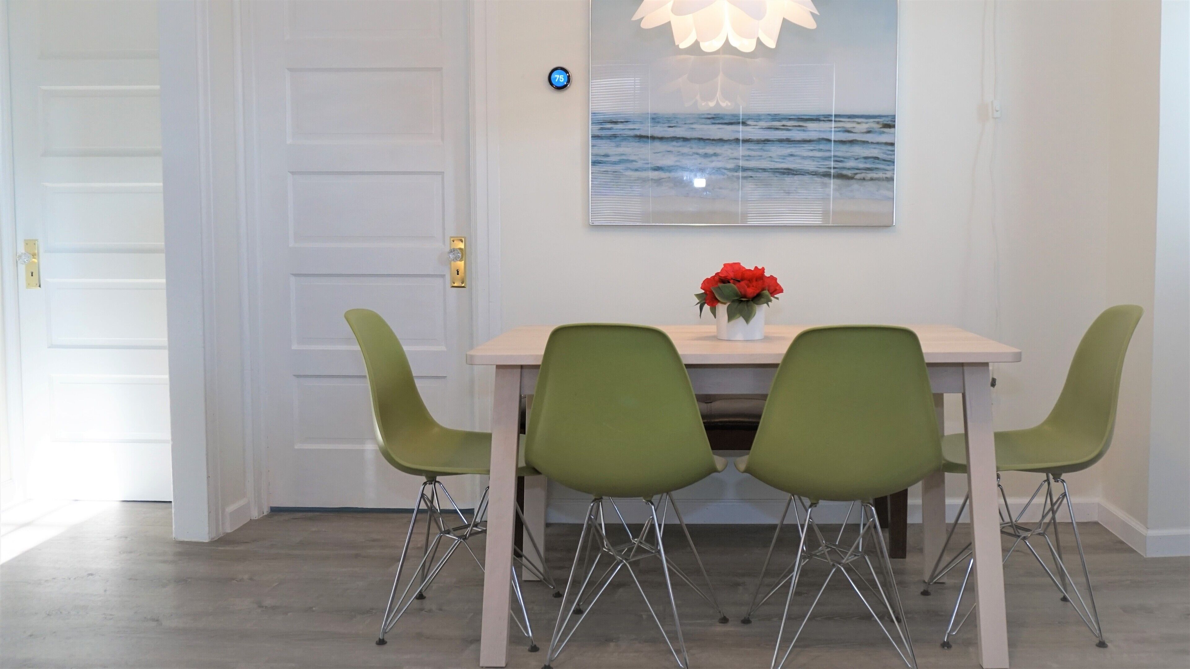 Un comedor con una mesa, cuatro sillas verdes, un jarrón con flores rojas y una obra de arte en la pared que representa el océano.