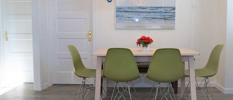 Un comedor con una mesa, cuatro sillas verdes, un jarrón con flores rojas y una obra de arte en la pared que representa el océano.