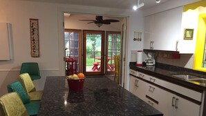 Una cocina con encimera de granito, un frutero con fruta roja y un comedor con sillas azul verdosas y amarillas.