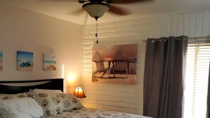 Una camera da letto con un letto, una ventola a soffitto, una lampada a parete e un quadro a tema spiaggia.