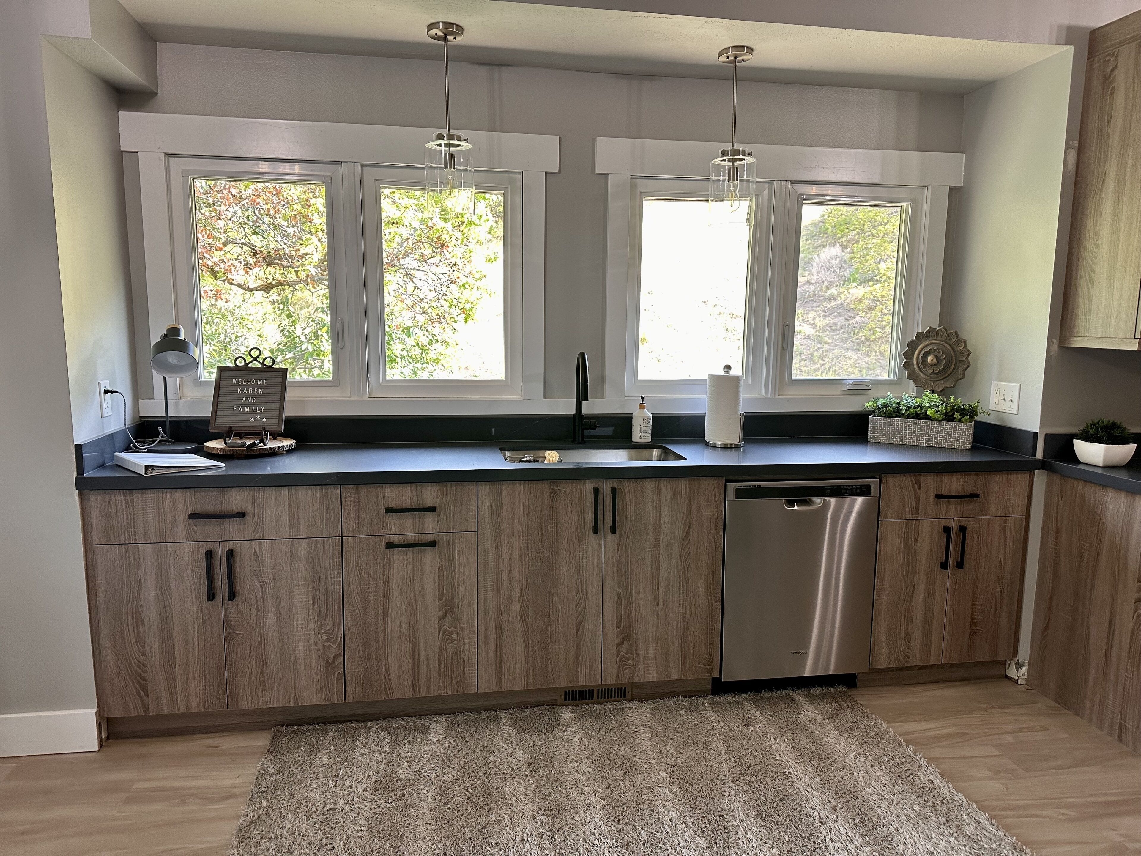 A modern kitchen with wooden cabinets, a stainless steel dishwasher, and a large window with a view of trees.