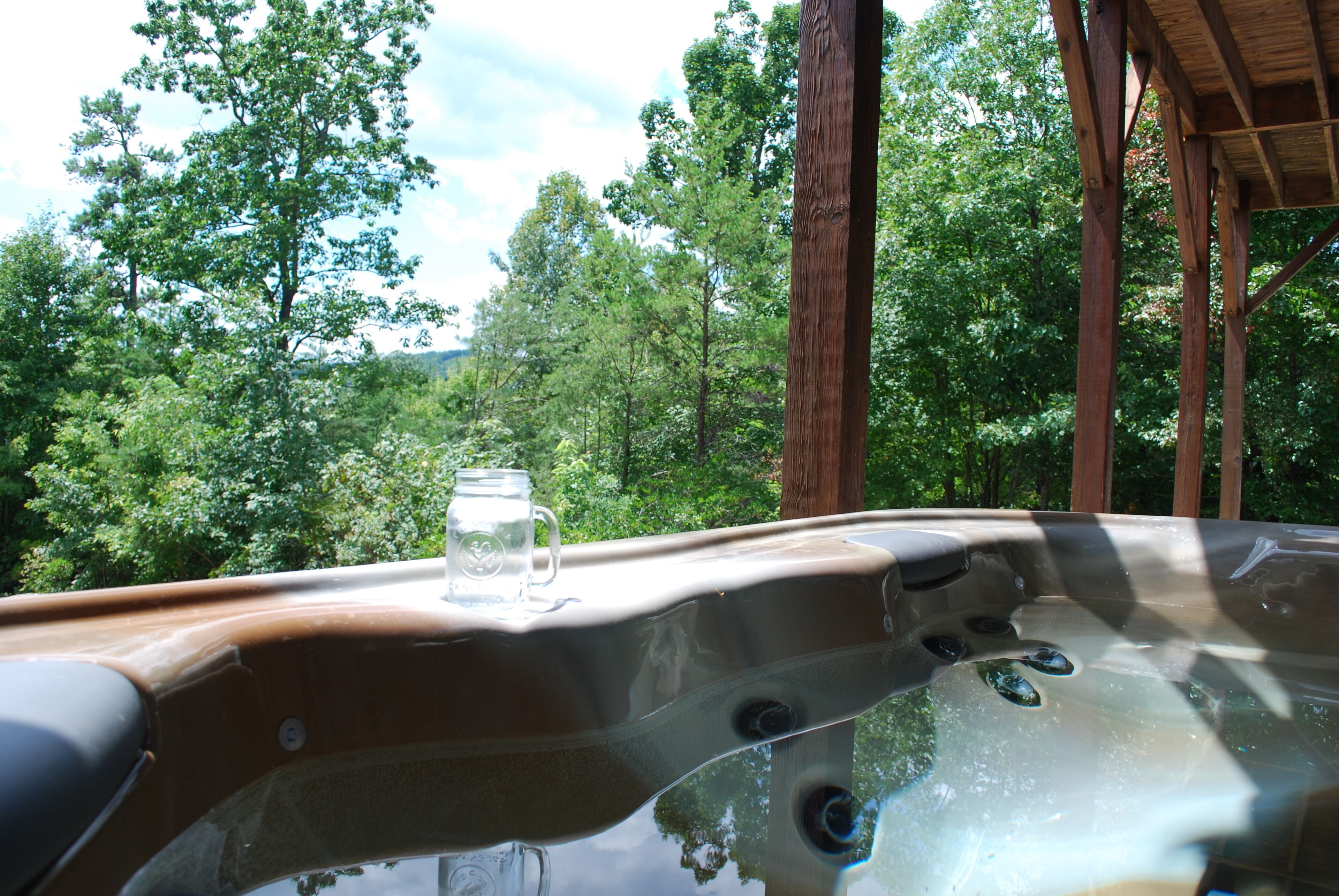 A hot tub with a glass of water and a mug, surrounded by a wooden deck and lush greenery.